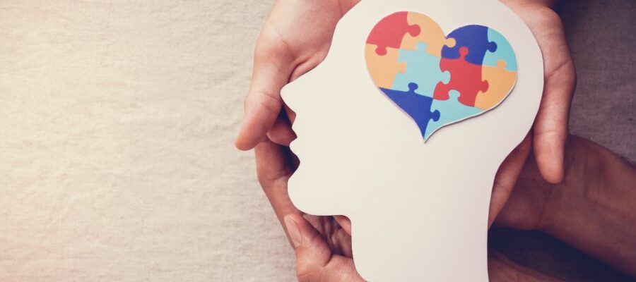 manos sosteniendo un carton con forma de cabeza humana con un puzzle en forma de corazón que hace referencia al cerebro